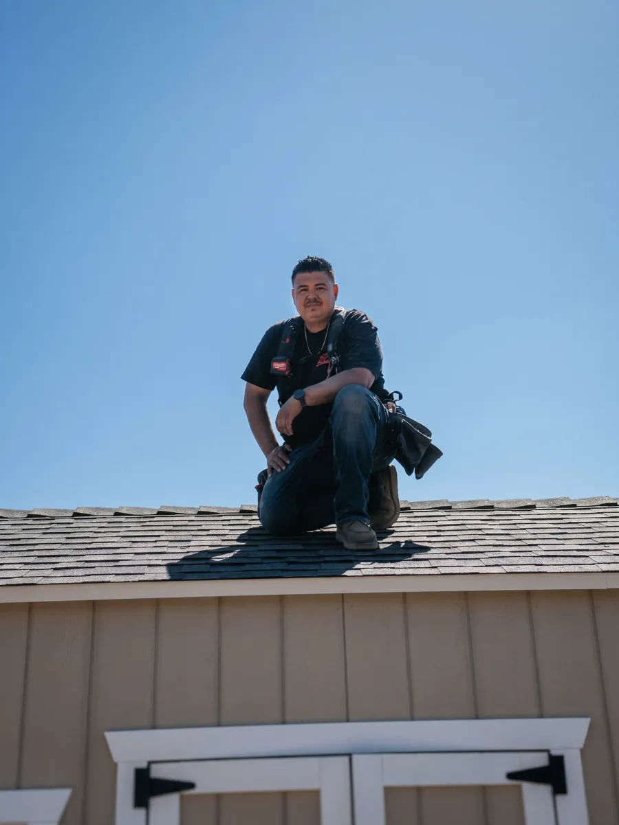 A 559Fences crew member kneeling on the roof of a finished shed against a clear sky, tool belt on.