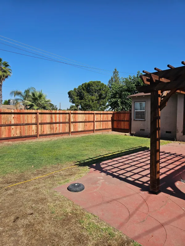 A finished wood fence enclosing a back yard with a pergola and a lawn.